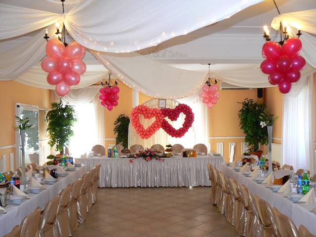 Uroczysta sala weselna udekorowana balonami w kształcie serc, zwisającymi z sufitu, oraz kwiatowymi kompozycjami na stołach nakrytych białymi obrusami.