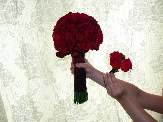 Dwa bukiety z ciemnoczerwonych róż, jeden duży trzymany w dłoni, drugi mały podawany, na tle zasłony w kwiatowy wzór.