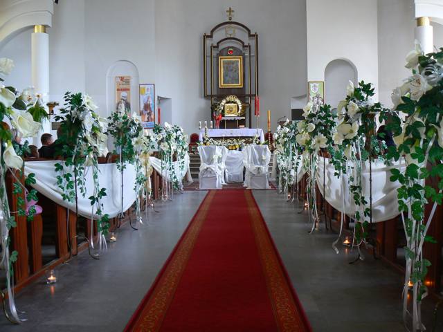 Uroczysta dekoracja kościoła przed ślubem: białe kwiaty, zielone pnącza i delikatne tkaniny zdobią ławki wzdłuż czerwonego dywanu prowadzącego do ołtarza. Świece w szklanych lampionach dodają...