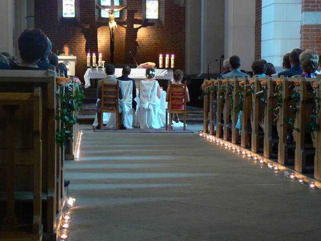 Dekoracja kościoła na ślub: rzędy ławek ozdobione pnączami zieleni z różami, ścieżka oświetlona małymi świeczkami, w tle siedząca para młoda podczas ceremonii.