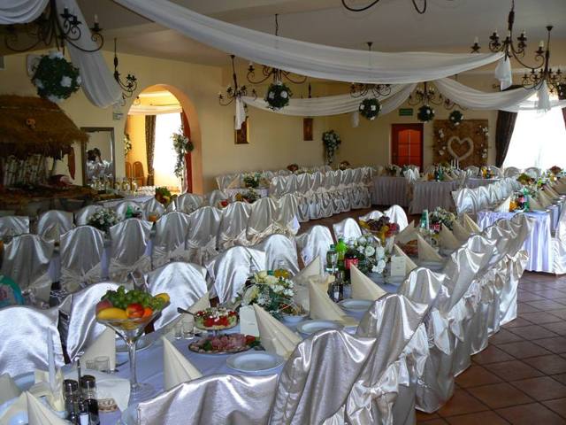 Sala weselna z długimi stołami udekorowanymi białymi obrusami i serwetkami, krzesła w białych pokrowcach z kokardami, dekoracje kwiatowe na stołach, owoce w szklanych wazach, białe drapowania...