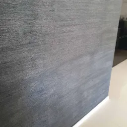 Szara ściana z wyraźną fakturą i delikatnym połyskiem, wykończona białą listwą przypodłogową, przylegająca do jasnej, błyszczącej podłogi.