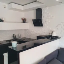 Nowoczesna kuchnia z czarnym blatem, białymi szafkami i ścianą z białej cegły, widoczny fragment sofy.