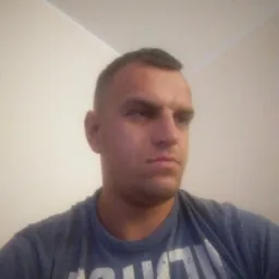 Mężczyzna w szarej koszulce z białym napisem, krótkie włosy, patrzy w prawo, neutralny wyraz twarzy, białe ściany w tle, amatorskie oświetlenie.