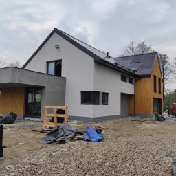MURZIK ANNA LEŚNIAK - Nowoczesny dom jednorodzinny z panelami słonecznymi na dachu, w trakcie wykończenia elewacji i zagospodarowania terenu, widoczny gruz i materiały budowlane, czerwony samochód dostawczy w tle.