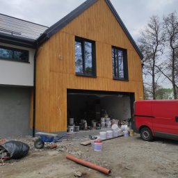 MURZIK ANNA LEŚNIAK - Nowoczesny dom z drewnianą elewacją w trakcie wykończenia, widoczne okna z czarnymi ramami, otwarty garaż z puszkami farb, czerwony van oraz nieporządek na placu budowy.