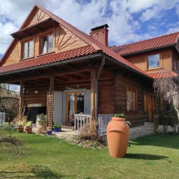 MURZIK ANNA LEŚNIAK - Dwupiętrowy dom z bali z czerwonym dachem i oknami, widok z ogrodu z glinianym zbiornikiem na deszczówkę.