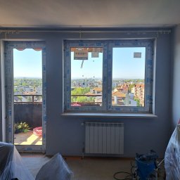 MURZIK ANNA LEŚNIAK - Nowe okna z widokiem na miasto i balkon, zabezpieczone folią meble w trakcie prac wykończeniowych, grzejnik pod oknem.