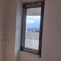 Szare okno z widokiem na miasto, zabezpieczone folią ochronną Aluplast, osadzone w surowej ścianie w trakcie remontu.