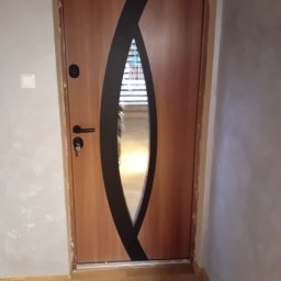 MURZIK Okna - Nowoczesne drzwi wejściowe z jasnego drewna z czarnym, owalnym zdobieniem, klamką i kluczem w zamku, w jasnym korytarzu.
