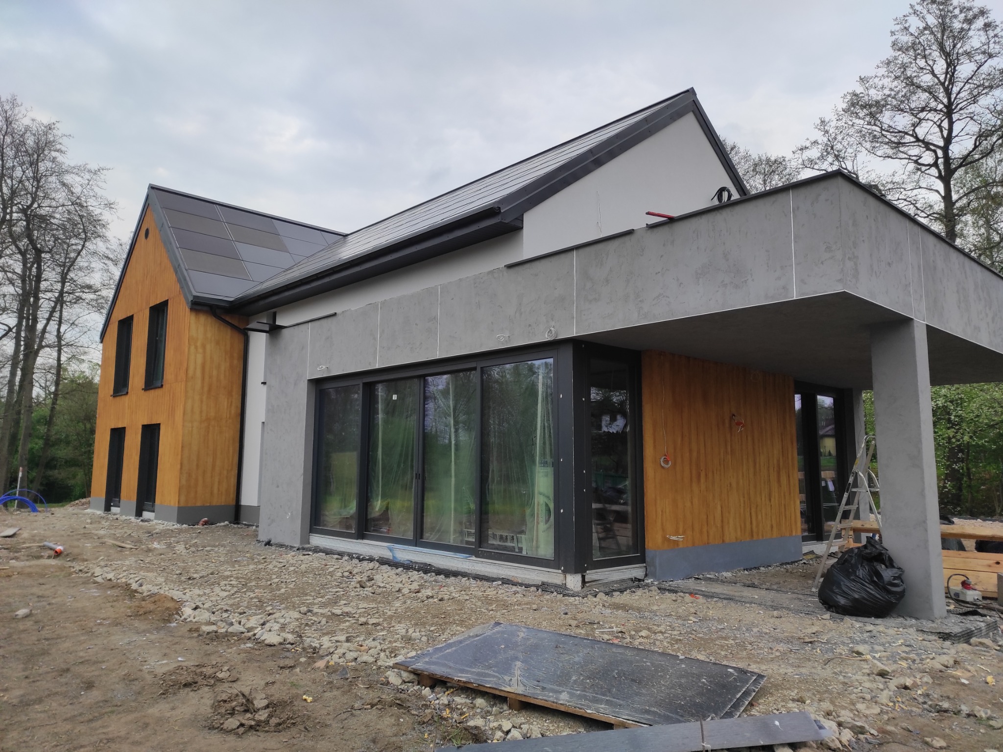Nowoczesny dom jednorodzinny w trakcie budowy, z dużymi przesuwnymi oknami, drewnianą elewacją i panelami słonecznymi na dachu, otoczony gruzem i materiałami budowlanymi.