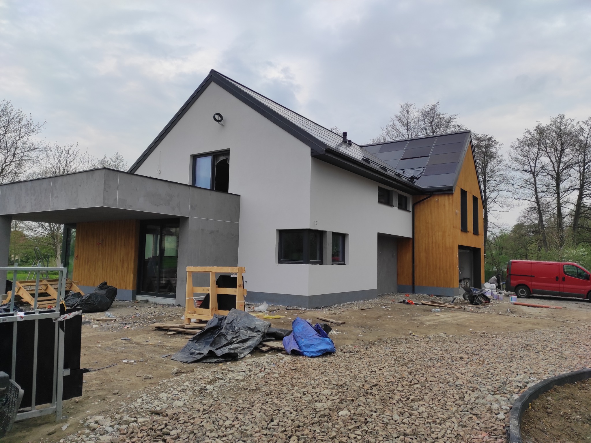 Nowoczesny dom jednorodzinny z panelami słonecznymi na dachu, w trakcie wykończenia elewacji i zagospodarowania terenu, widoczny gruz i materiały budowlane, czerwony samochód dostawczy w tle.