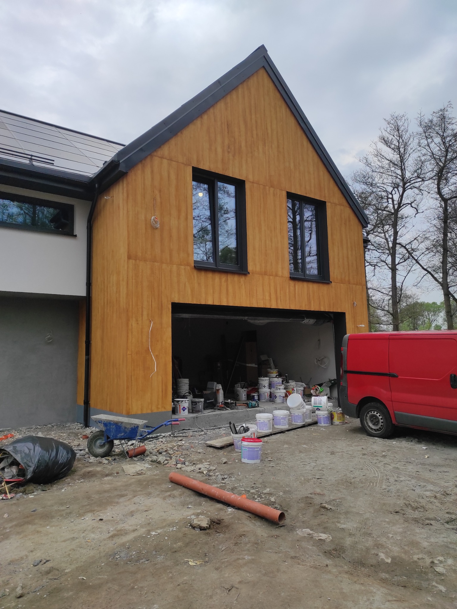 Nowoczesny dom z drewnianą elewacją w trakcie wykończenia, widoczne okna z czarnymi ramami, otwarty garaż z puszkami farb, czerwony van oraz nieporządek na placu budowy.