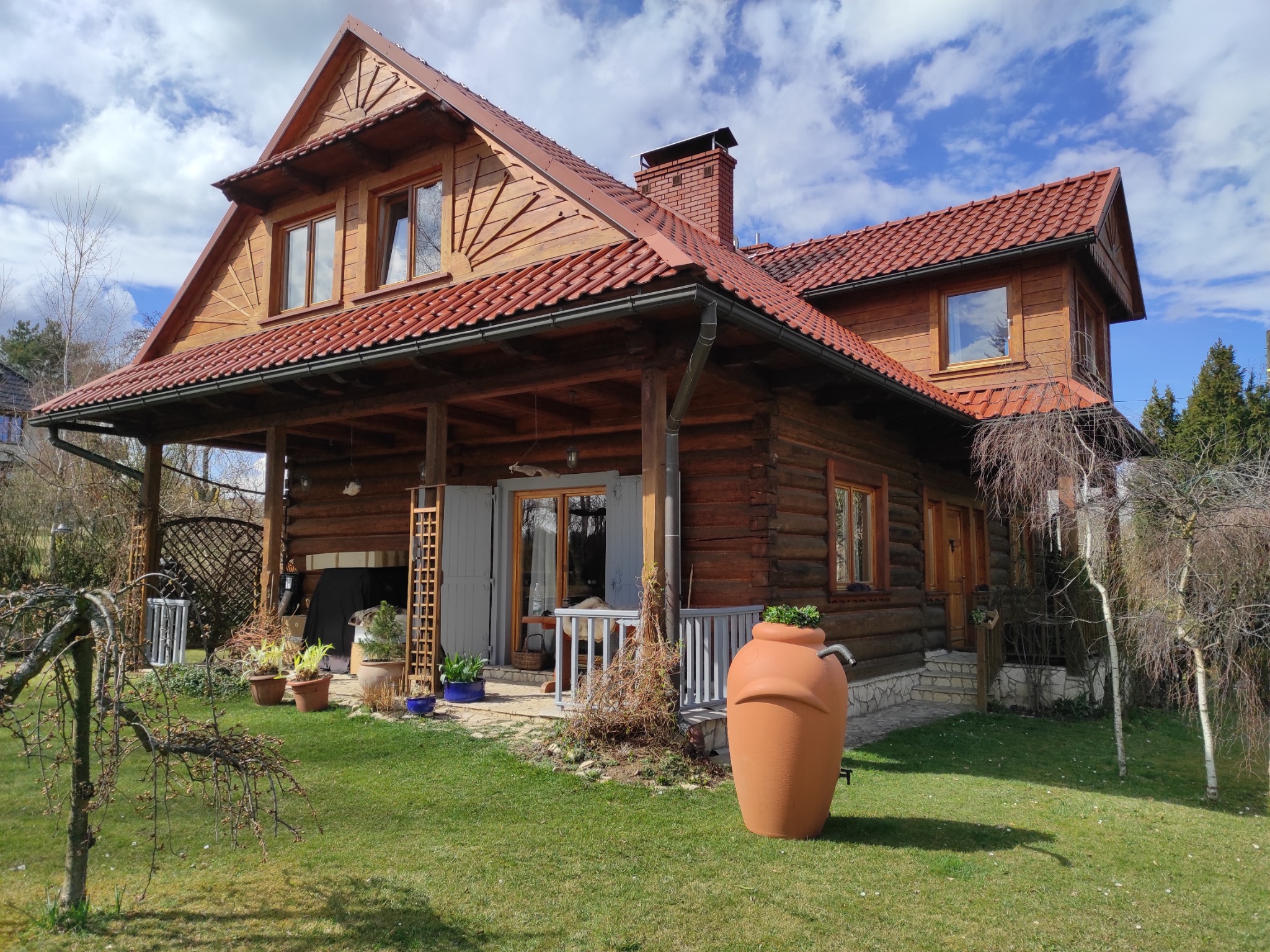 Dwupiętrowy dom z bali z czerwonym dachem i oknami, widok z ogrodu z glinianym zbiornikiem na deszczówkę.