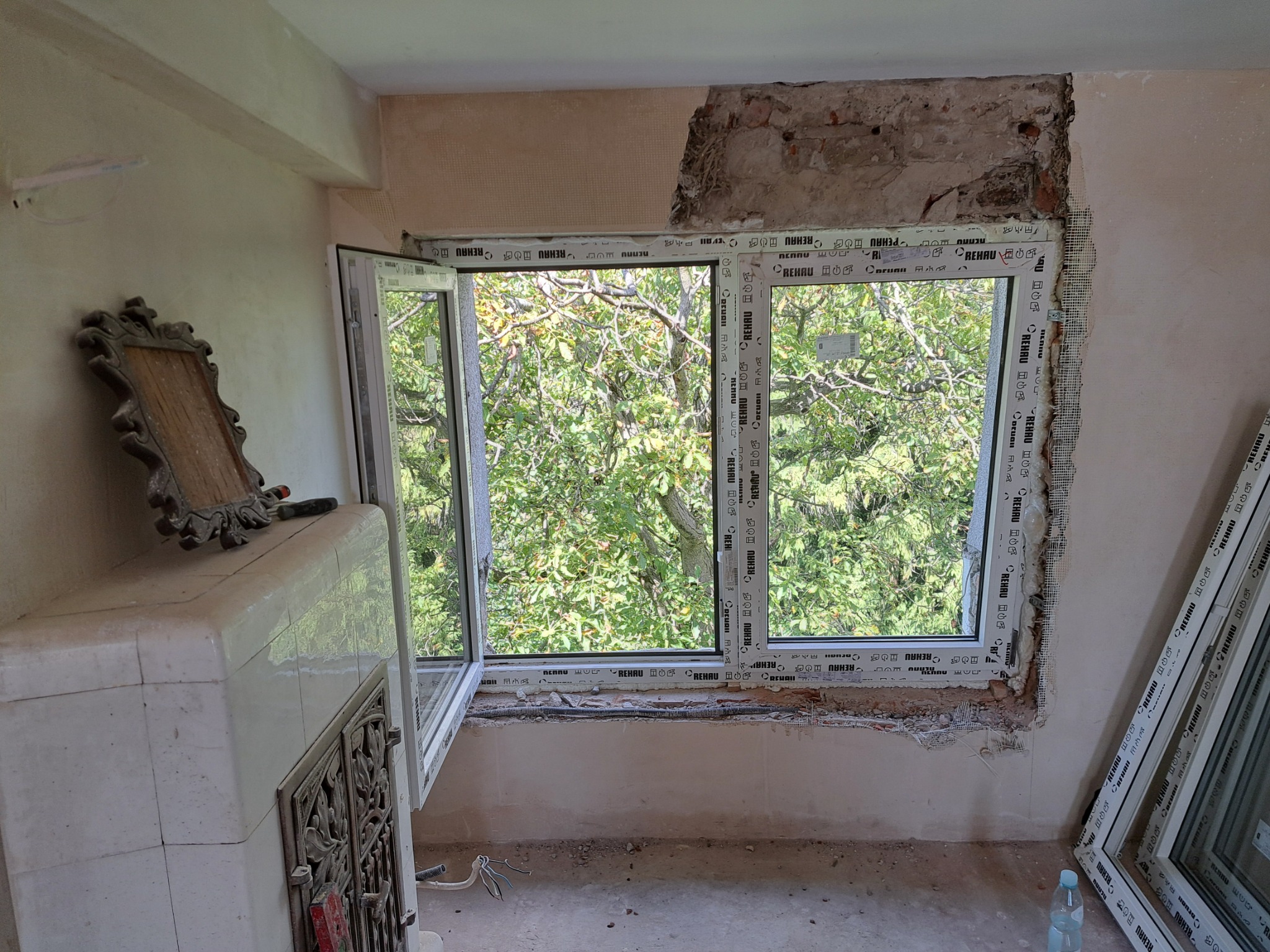 Montaż okna PCV w trakcie remontu: widoczne surowe ściany, fragment starego okna, nowe okno częściowo zamontowane z widokiem na zieleń, oraz elementy wykończeniowe oparte o ścianę; w tle piec...