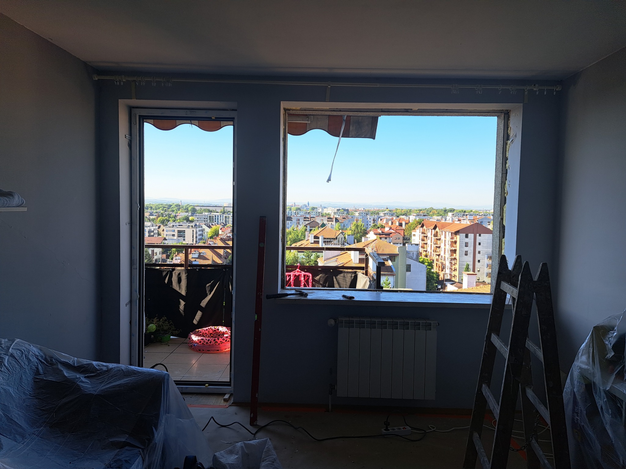 Wnętrze pokoju w trakcie remontu z otwartym oknem i drzwiami balkonowymi, widok na miasto, grzejnik, drabina i narzędzia widoczne.