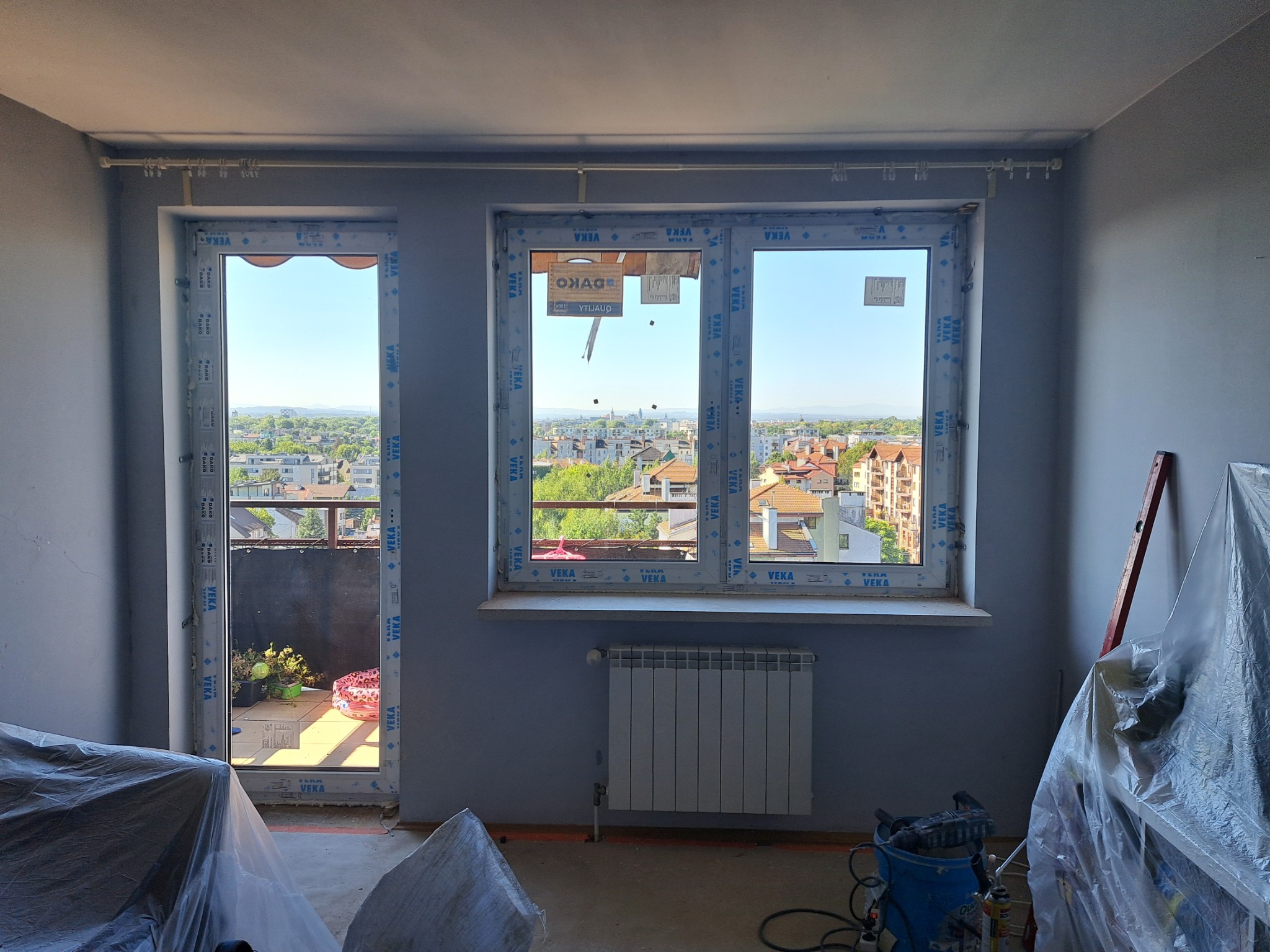Nowe okna z widokiem na miasto i balkon, zabezpieczone folią meble w trakcie prac wykończeniowych, grzejnik pod oknem.