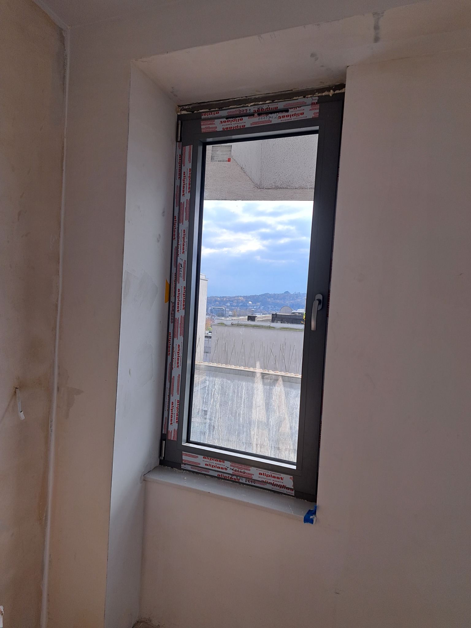 Szare okno z widokiem na miasto, zabezpieczone folią ochronną Aluplast, osadzone w surowej ścianie w trakcie remontu.