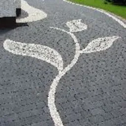 Ułożona z ciemnoszarej kostki brukowej ścieżka z dekoracyjnym wzorem kwiatowym z jasnych kamieni, prowadząca wzdłuż ściany budynku i dalej przez ogród.