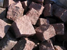 Stos nieregularnych kostek granitowych w odcieniach czerwieni i brązu, o szorstkiej fakturze, oświetlonych nierównomiernym światłem słonecznym podkreślającym ich naturalny wygląd.