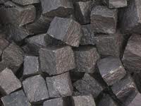 Stos szarych, surowych kostek brukowych o nieregularnych kształtach i chropowatej fakturze, ułożonych blisko siebie.