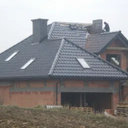 Budowa domu z cegły: czarny dach z oknami dachowymi, widoczny komin i pracownik na dachu. Częściowo wykończona elewacja, palety na pierwszym planie.