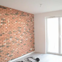 Ściana w pokoju wykończona dekoracyjną cegłą w odcieniach czerwieni, szarości i bieli, z widocznymi przewodami oświetleniowymi na podłodze i białym oknem balkonowym w tle.