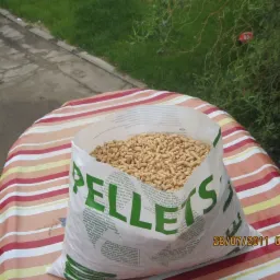 Otwarty worek z granulatem opałowym na stole w paski, widoczna data i godzina na worku, w tle trawa i chodnik.