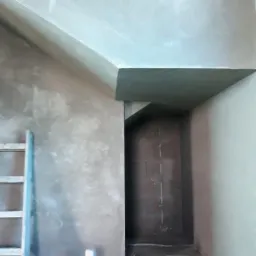 Wnętrze w trakcie prac wykończeniowych: ściany pokryte świeżym tynkiem, widoczna drabina oparta o ścianę, narzędzia na podłodze i fragment sufitu z nietypowym załamaniem.