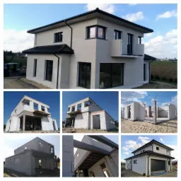 Dwukondygnacyjny dom w stanie surowym zamkniętym, z szarą elewacją, czarną dachówką i stolarką okienną, widoczny z zewnątrz na tle błękitnego nieba i zieleni.