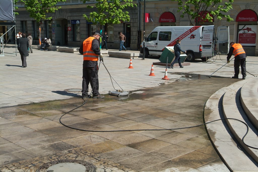 Pracownik w pomarańczowej kamizelce czyści kostkę brukową na placu za pomocą myjki ciśnieniowej, widoczny efekt czyszczenia i brudna woda, w tle biały samochód dostawczy i budynki miejskie.
