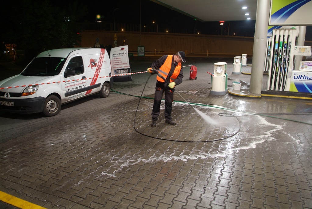 Pracownik w kamizelce odblaskowej czyści kostkę brukową na stacji benzynowej za pomocą myjki ciśnieniowej, widoczny biały samochód dostawczy z logo firmy i otwarte drzwi boczne.