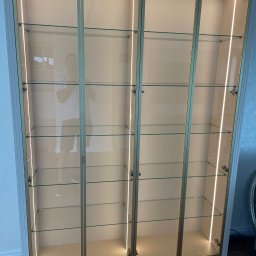 PAPA - MEBLE RAFAŁ ŻELAZNY - Wysoka szafa wnękowa z przeszklonymi drzwiami, podświetlona listwami LED, z widocznym odbiciem osoby w drzwiach.