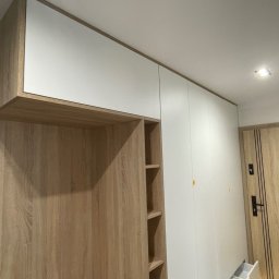 PAPA - MEBLE RAFAŁ ŻELAZNY - Jasna szafa wnękowa z drewnianymi elementami i białymi frontami, widoczne półki w otwartej części, obok drzwi wejściowe.