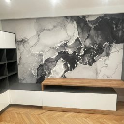 PAPA - MEBLE RAFAŁ ŻELAZNY - Nowoczesna szafka RTV z białymi frontami, drewnianym blatem i otwartymi półkami na tle ściany z abstrakcyjnym wzorem w odcieniach szarości.