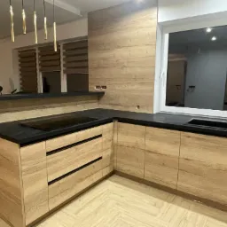 Nowoczesna kuchnia z frontami z drewna w odcieniach beżu i czarnym blatem, oświetlona wiszącymi lampami w złotym kolorze, z widocznym oknem.