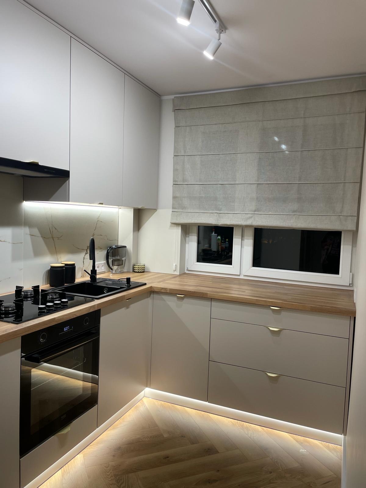 Nowoczesna kuchnia w odcieniach szarości z drewnianym blatem, czarnym zlewem i płytą grzewczą, oświetleniem LED pod szafkami i roletą rzymską w oknie.