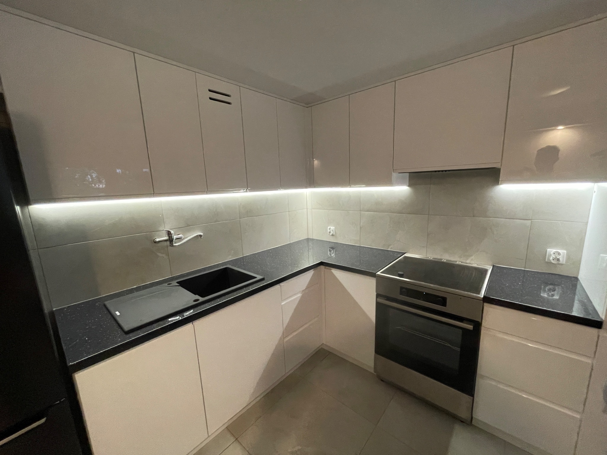 Nowoczesna kuchnia z białymi, lakierowanymi szafkami, czarnym, błyszczącym blatem i podświetleniem LED pod szafkami wiszącymi.