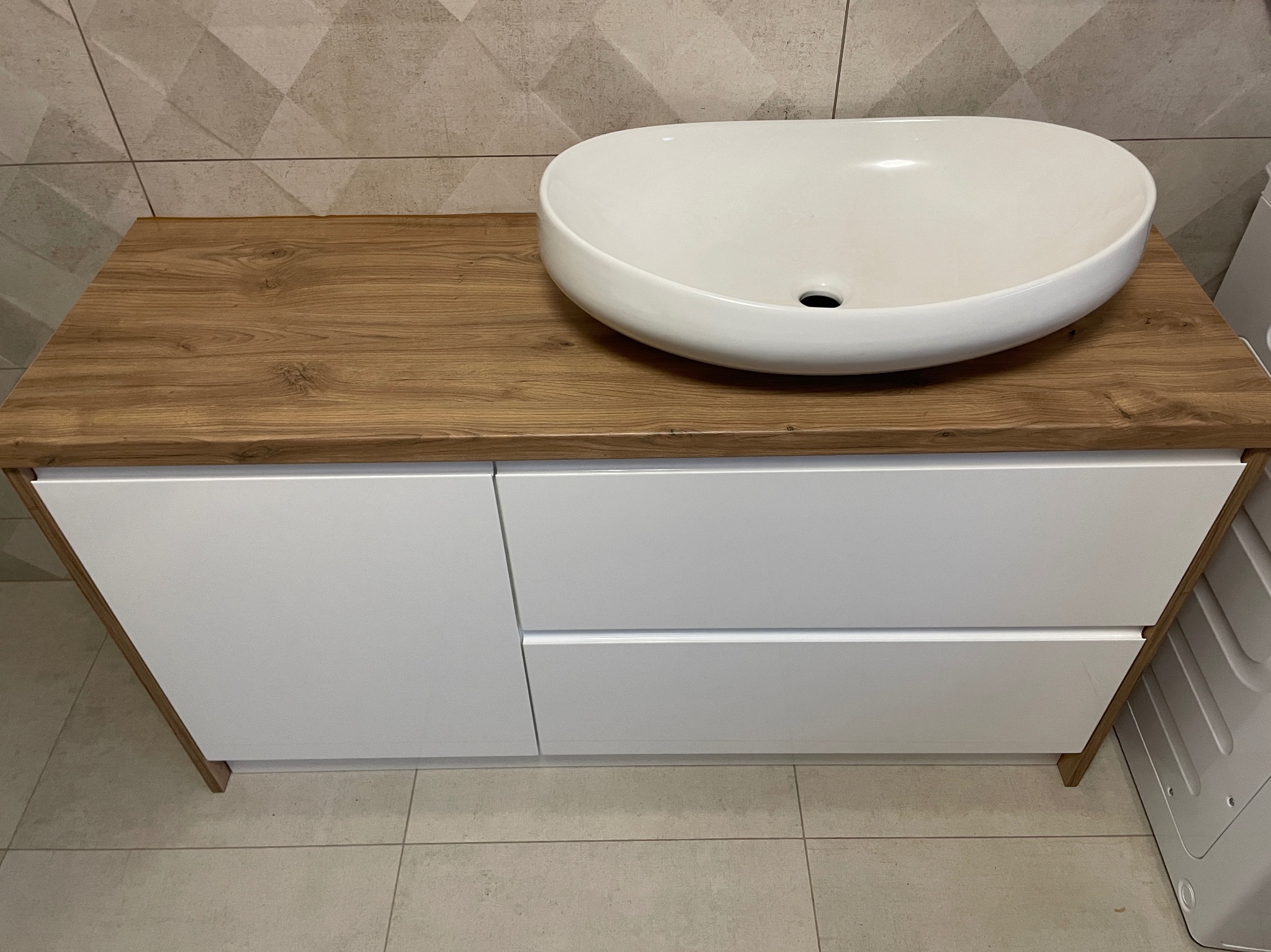 Biała, owalna umywalka nablatowa na drewnianym blacie z białymi szafkami w łazience z geometrycznymi płytkami na ścianie.