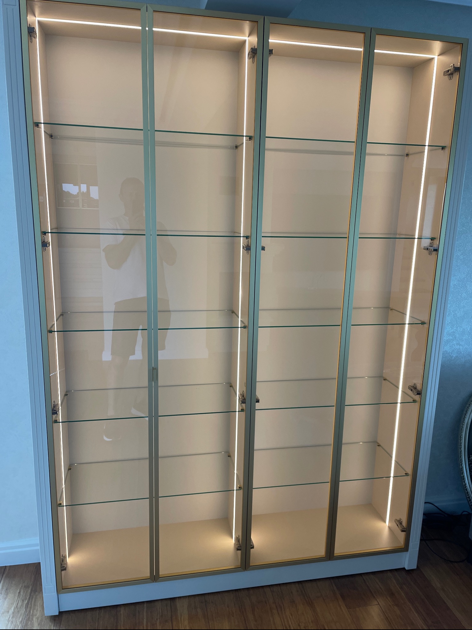 Wysoka szafa wnękowa z przeszklonymi drzwiami, podświetlona listwami LED, z widocznym odbiciem osoby w drzwiach.