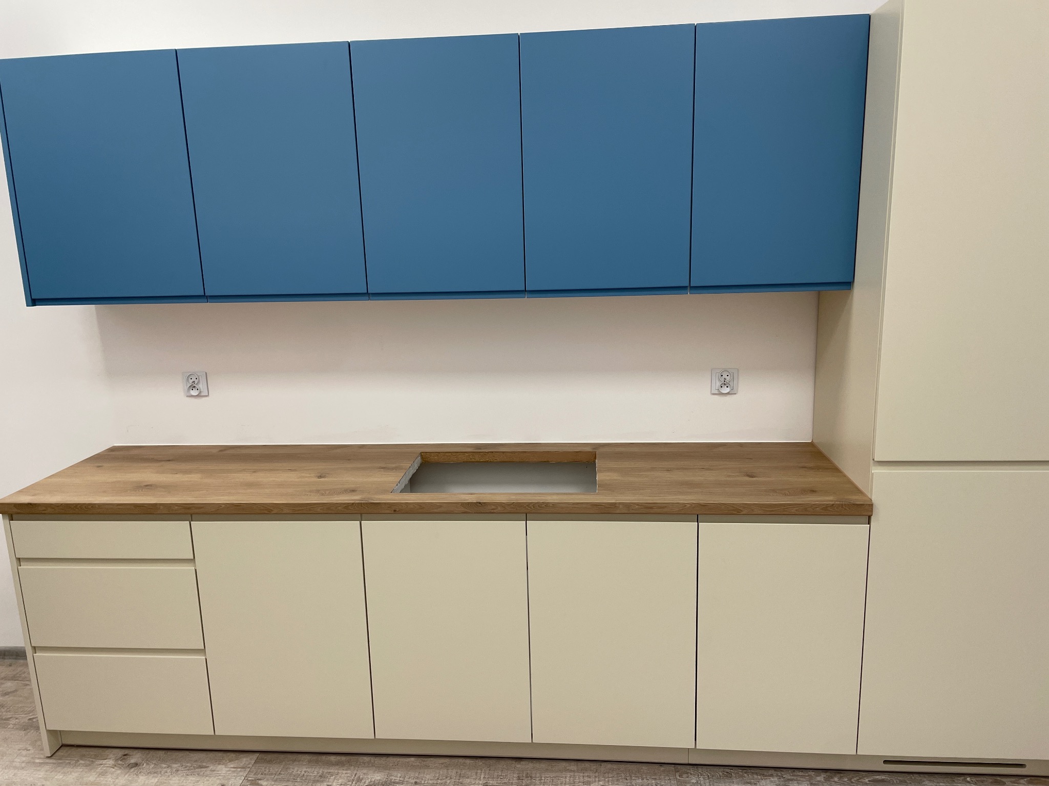 Nowoczesna zabudowa kuchenna z drewnianym blatem z wyciętym otworem na zlew, górne szafki w kolorze niebieskim, dolne i wysoka szafka w kolorze kremowym, na białej ścianie widoczne dwa gniazdka...