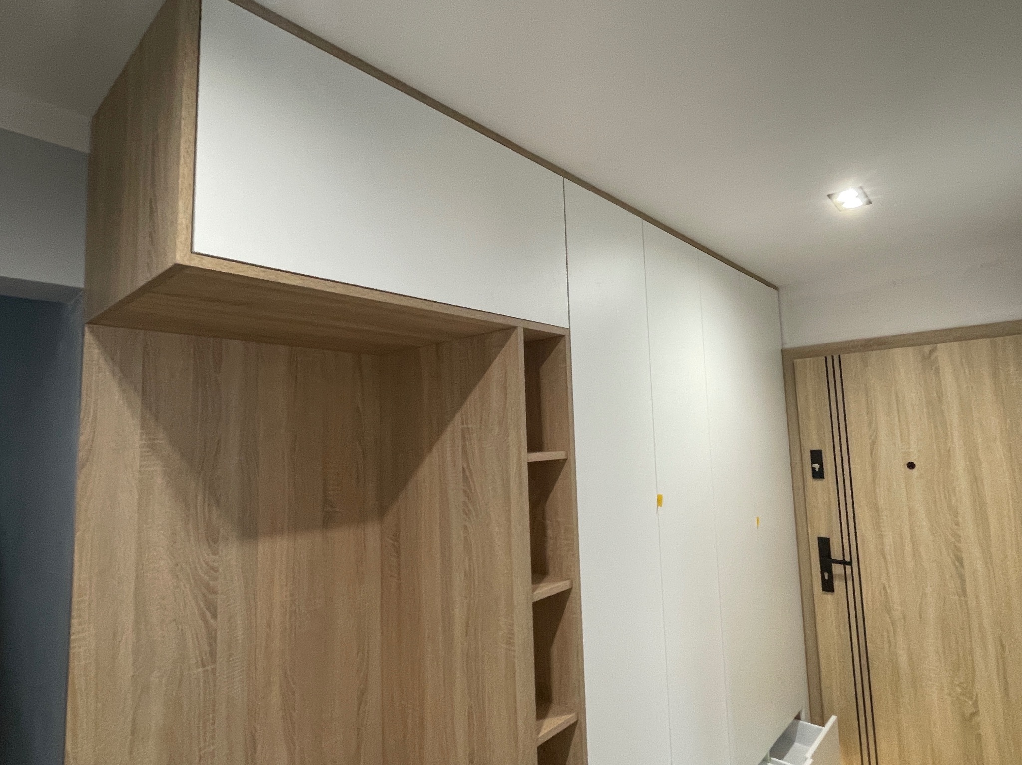 Jasna szafa wnękowa z drewnianymi elementami i białymi frontami, widoczne półki w otwartej części, obok drzwi wejściowe.