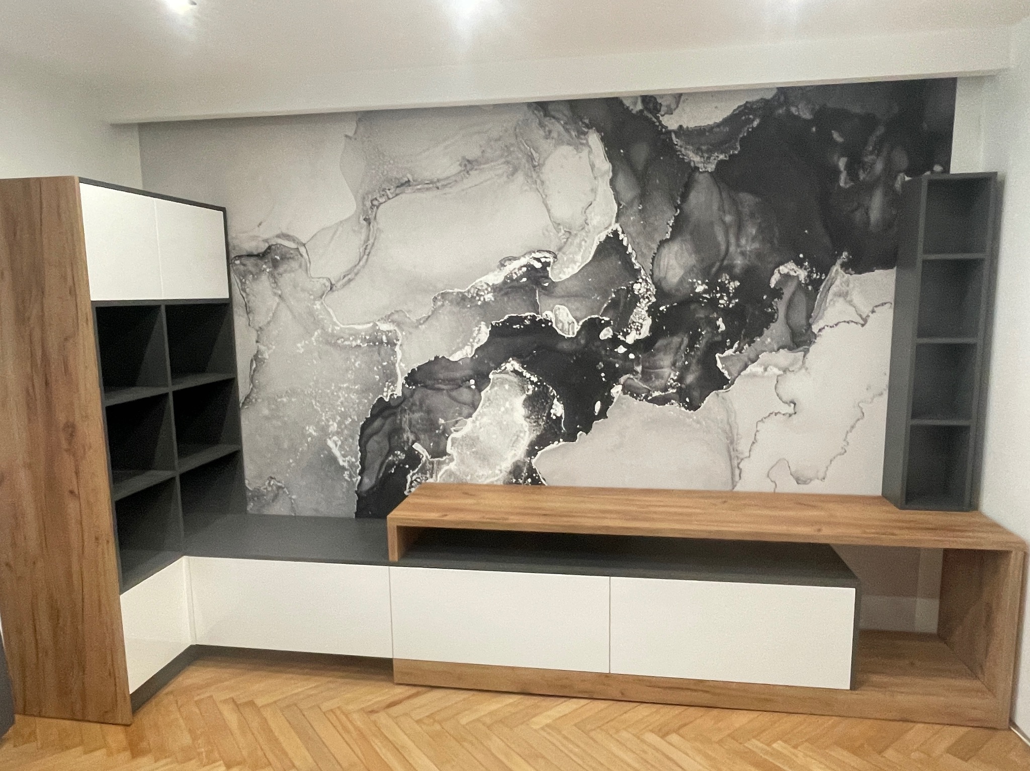 Nowoczesna szafka RTV z białymi frontami, drewnianym blatem i otwartymi półkami na tle ściany z abstrakcyjnym wzorem w odcieniach szarości.
