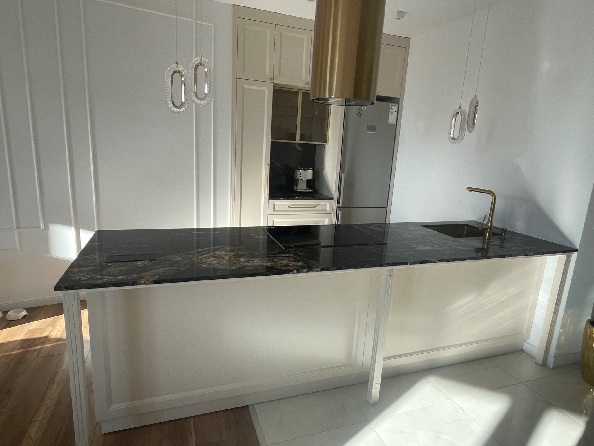 Nowoczesna kuchnia z wyspą i blatem z ciemnego granitu, złota armatura i okap, kremowe szafki z frezowanymi frontami, wiszące lampy o owalnym kształcie.