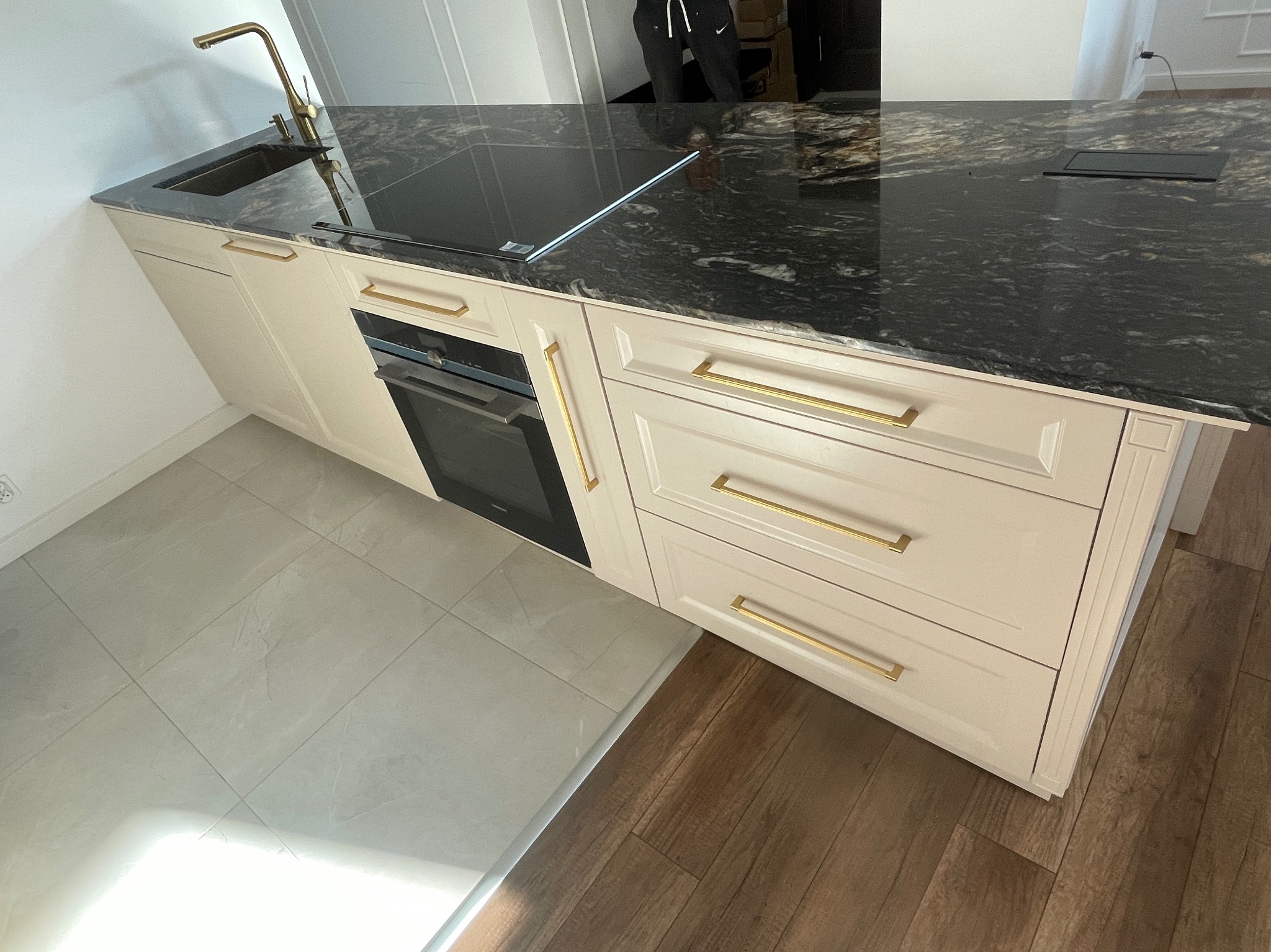 Fragment kuchni z beżowymi szafkami z frezowanymi frontami i złotymi uchwytami, czarny blat z marmurkowym wzorem, złota bateria kuchenna.