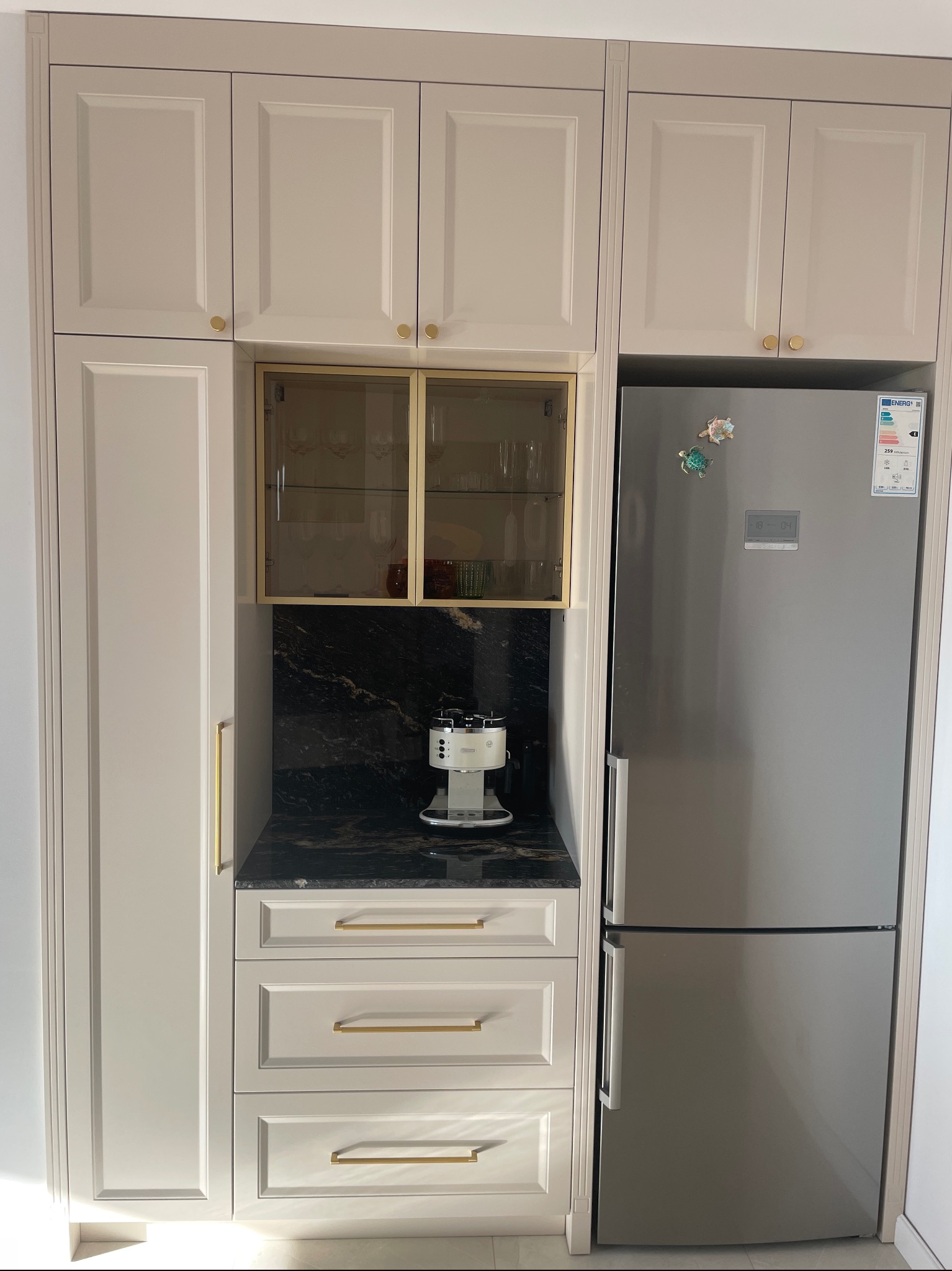 Zabudowa kuchenna w odcieniach beżu z czarnym blatem, złotymi uchwytami i przeszkloną szafką, obok stoi lodówka z magnesami.