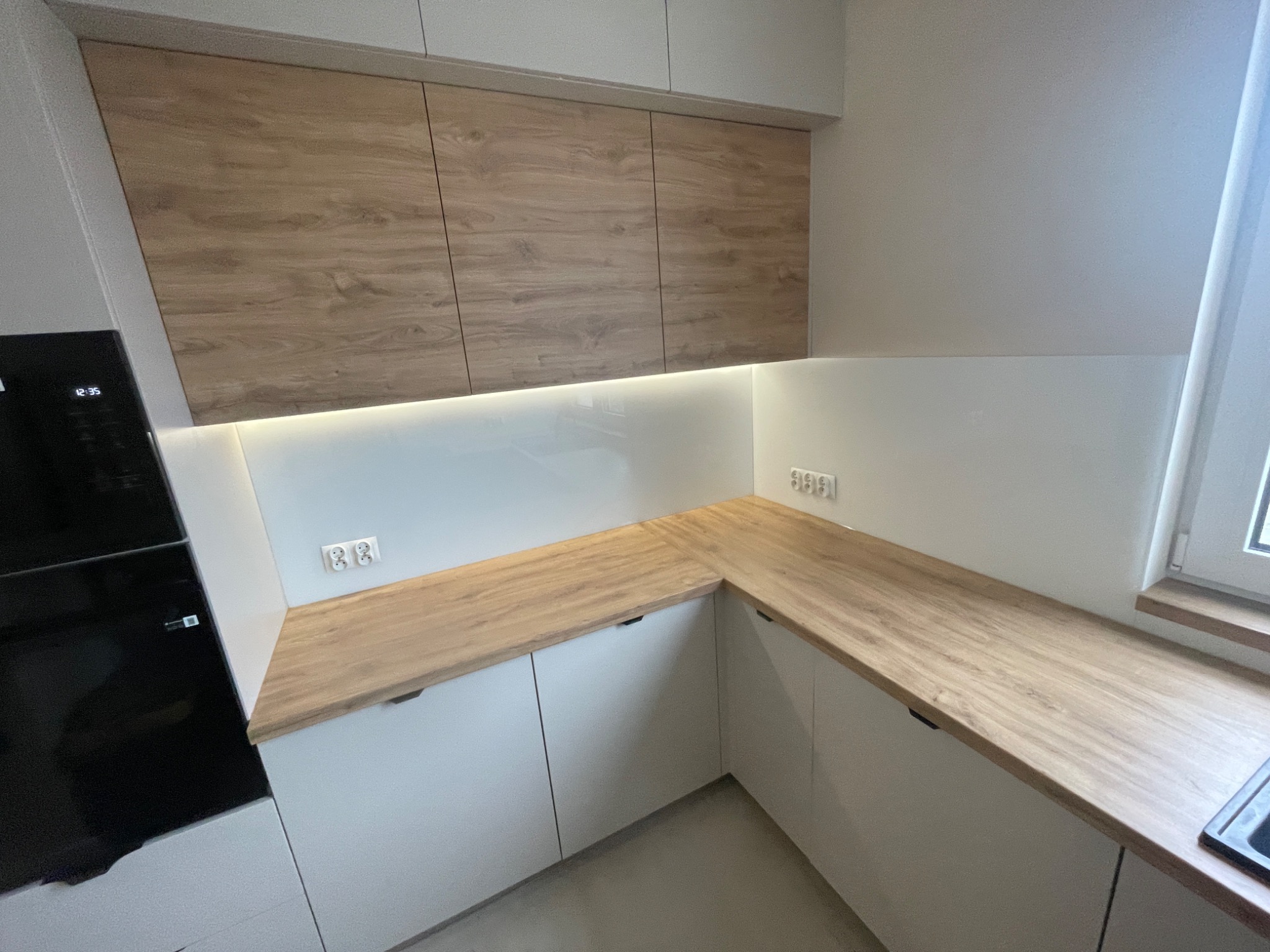 Nowoczesna kuchnia z białymi szafkami dolnymi i górnymi wykończonymi drewnopodobnym blatem, oświetlenie LED podszafkowe, widoczny fragment okna i piekarnika.