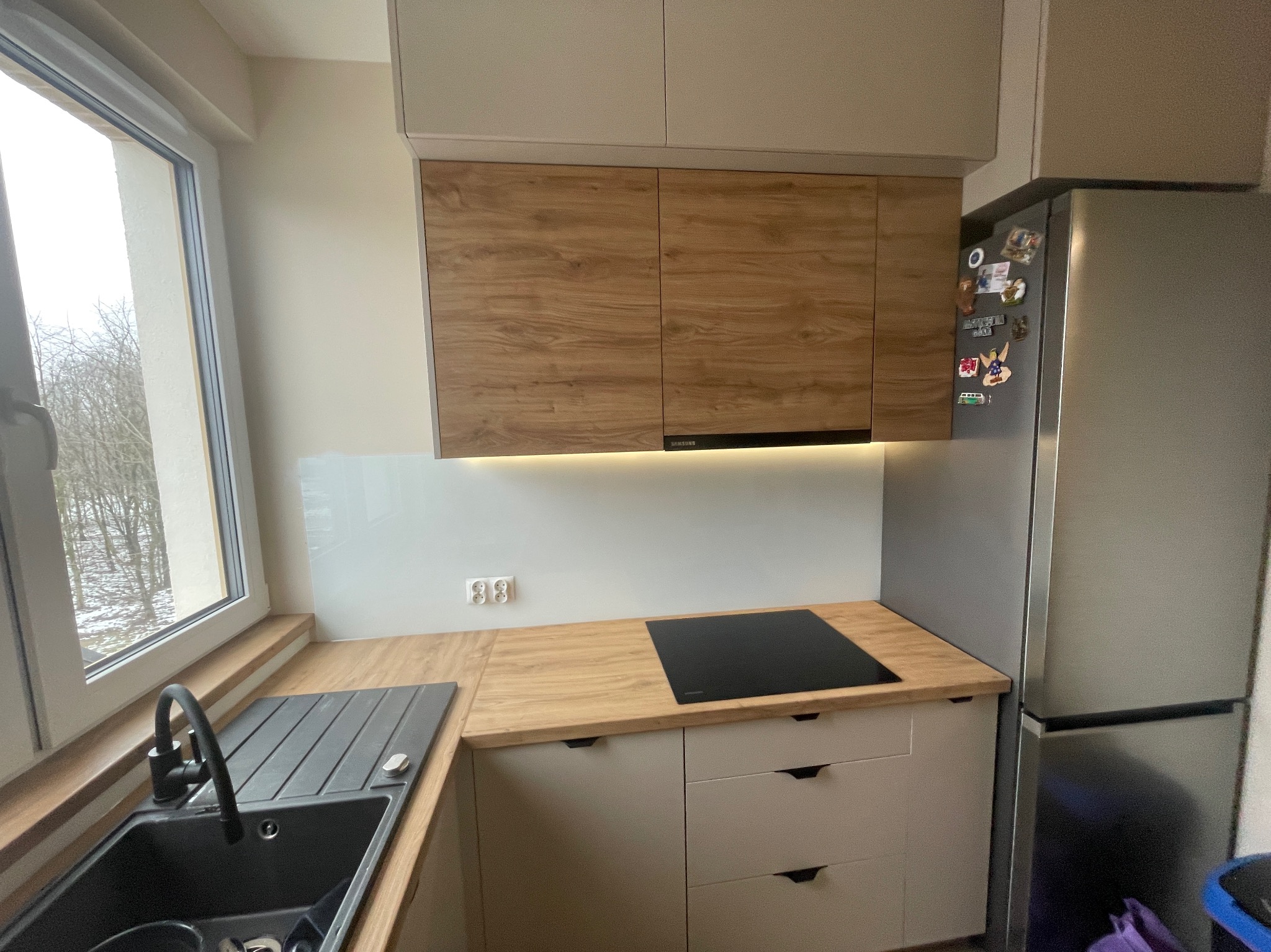 Nowoczesna kuchnia z drewnianym blatem, indukcyjną płytą grzewczą, jasnymi szafkami i widokiem na zimowy krajobraz za oknem.