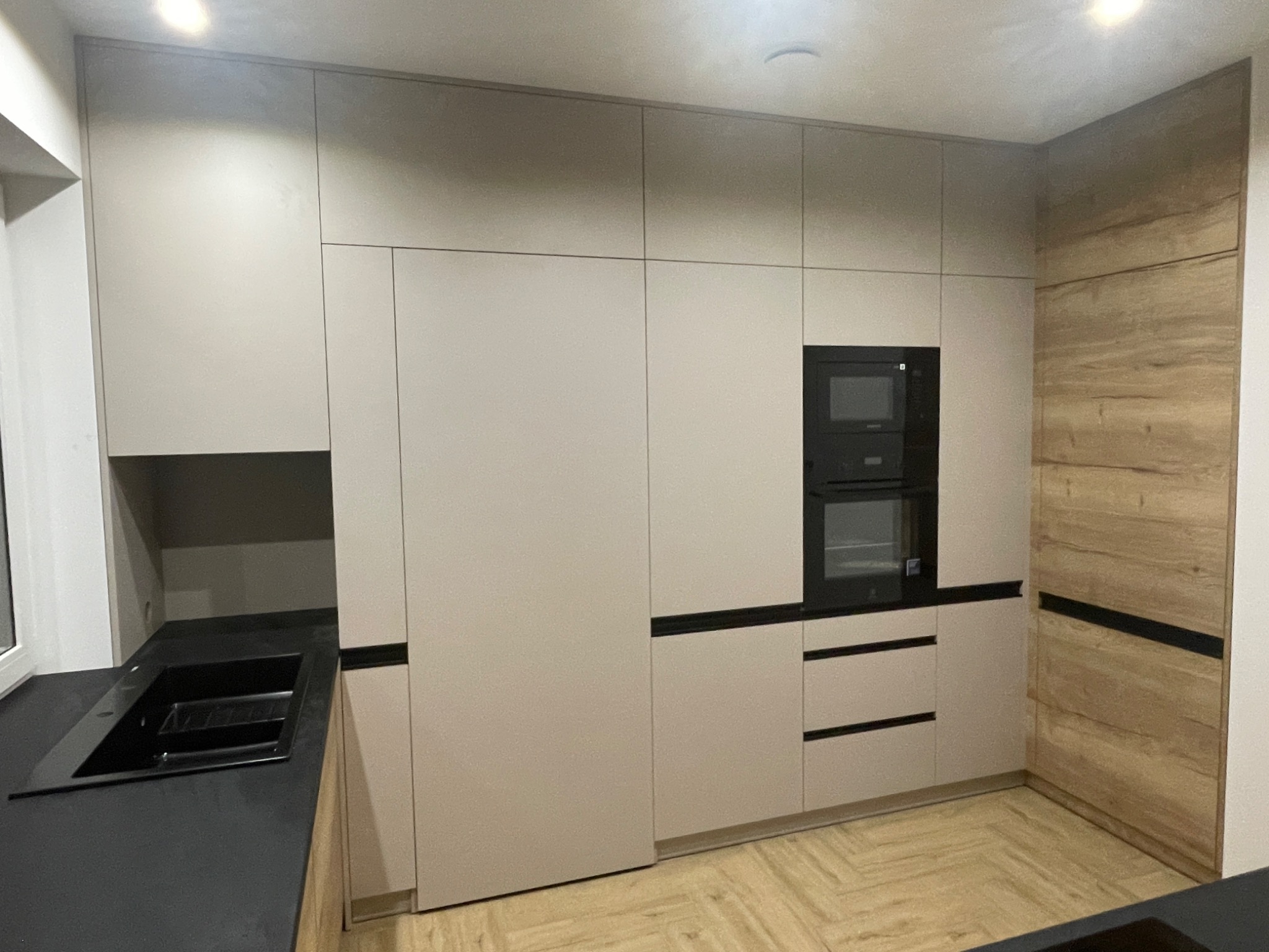 Nowoczesna kuchnia z zabudową w odcieniach beżu i drewna, z czarnym blatem i wbudowanym piekarnikiem i mikrofalówką.