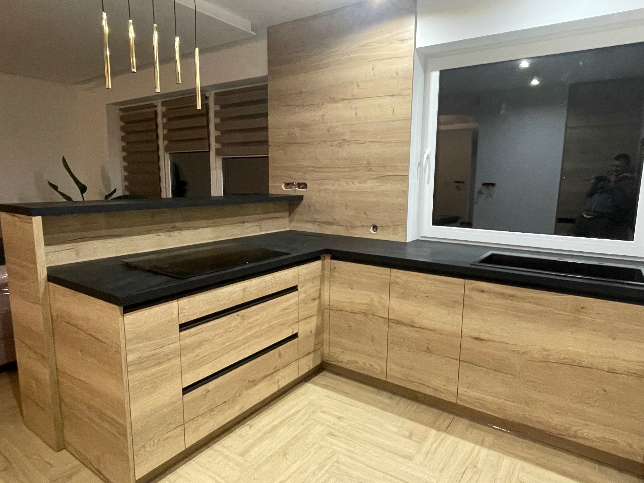 Nowoczesna kuchnia z frontami z drewna w odcieniach beżu i czarnym blatem, oświetlona wiszącymi lampami w złotym kolorze, z widocznym oknem.